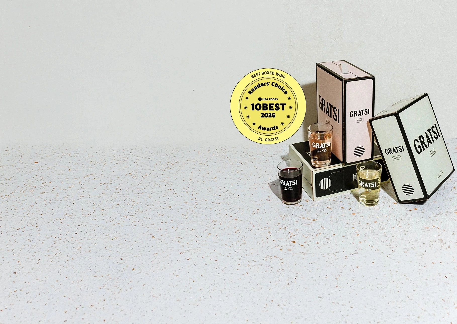 Three wine glasses, three Gratsi boxed wines, and a yellow 10Best 2026 Best Boxed Wine award on a light speckled surface against a plain background.