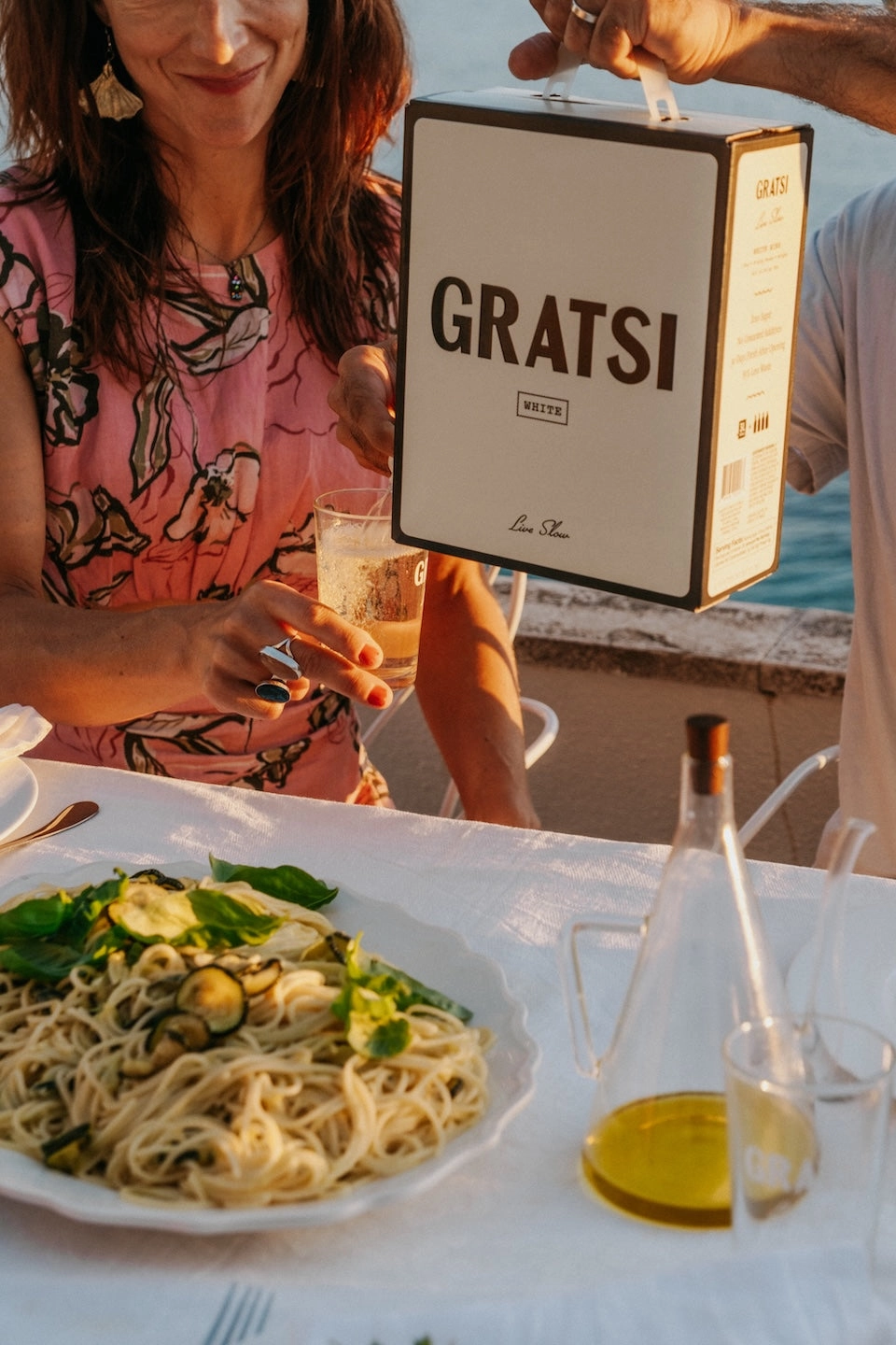 A person pours white wine from a boxed Gratsi container into a glass for a woman at an outdoor table set with pasta, olive oil, and basil, overlooking water.