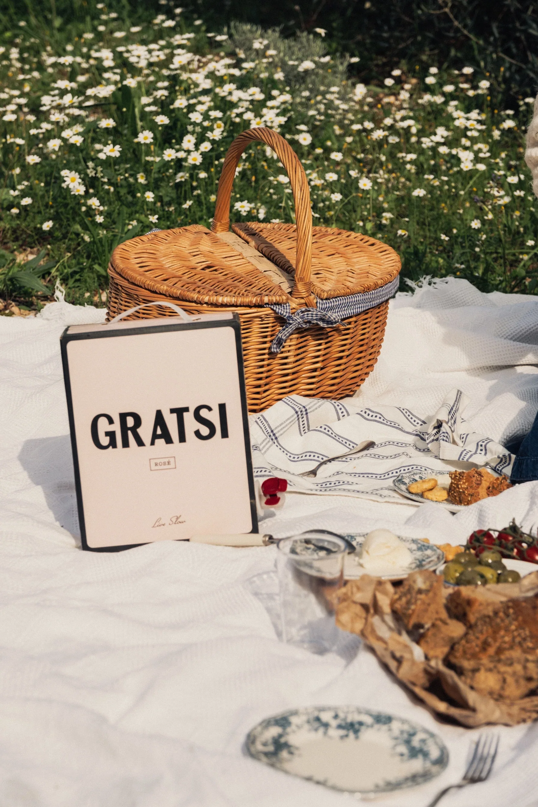 A picnic setup on a white blanket features a wicker basket, a menu card reading Gratsi, plates with food, grapes, cheese, bread, and flowers in the background.