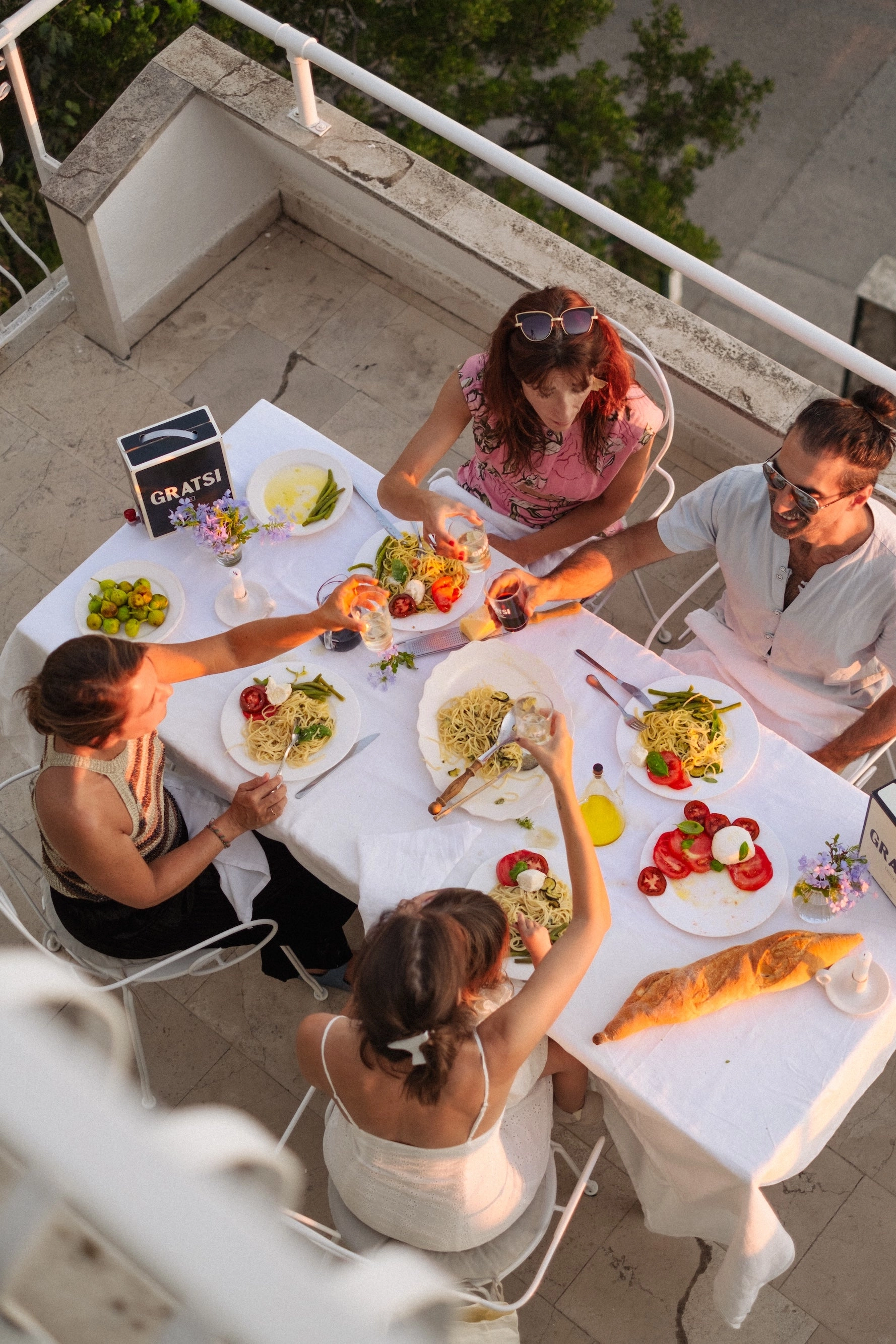 Four people sit around a table outdoors, sharing a meal with pasta, bread, salads, and drinks. The atmosphere is warm and friendly as they toast together. The table is decorated with small flowers and a Gratsi book.