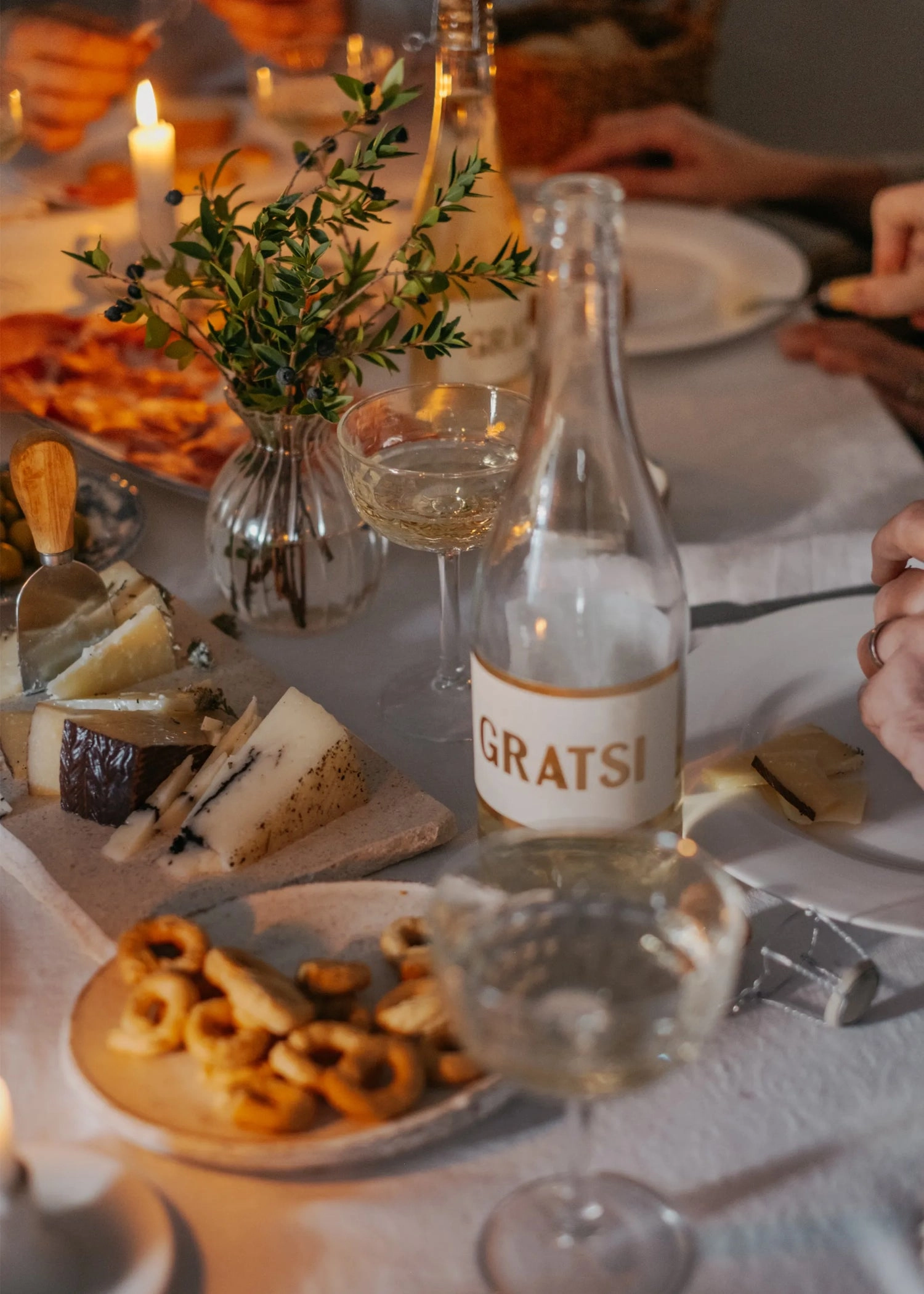 A table set for a meal features a bottle labeled Gratsi, filled wine glasses, a cheese platter with various cheeses, crackers, a small vase with greenery, and lit candles, creating a cozy, inviting atmosphere.