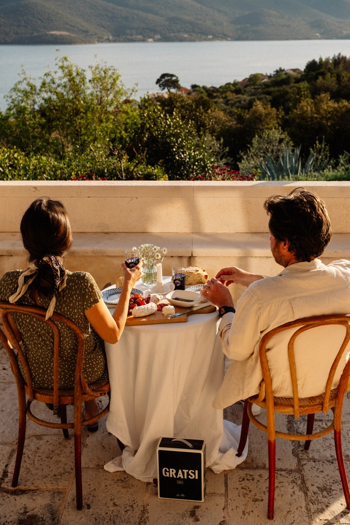 A man and woman sit at a small outdoor table set with wine, cheese, and snacks, overlooking a scenic view of water and forested hills. A black bag labeled Gratsi rests on the stone floor by their chairs.