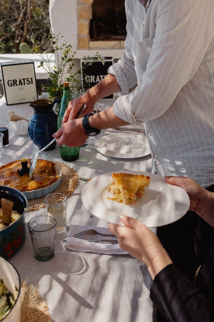 A person serves a slice of savory pie onto a plate at a sunlit outdoor table set with dishes, glasses, and flowers, while another person holds the plate. Gratsi signs are visible in the background.