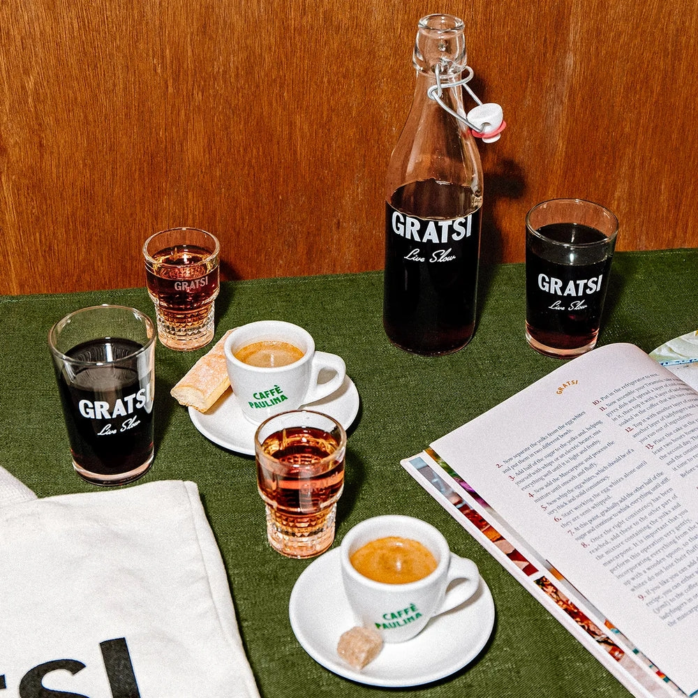 Three espresso cups with saucers, sugar cubes, and biscotti sit on a green tablecloth, surrounded by glasses and a bottle labeled Gratsi, alongside an open book and a white tote bag.