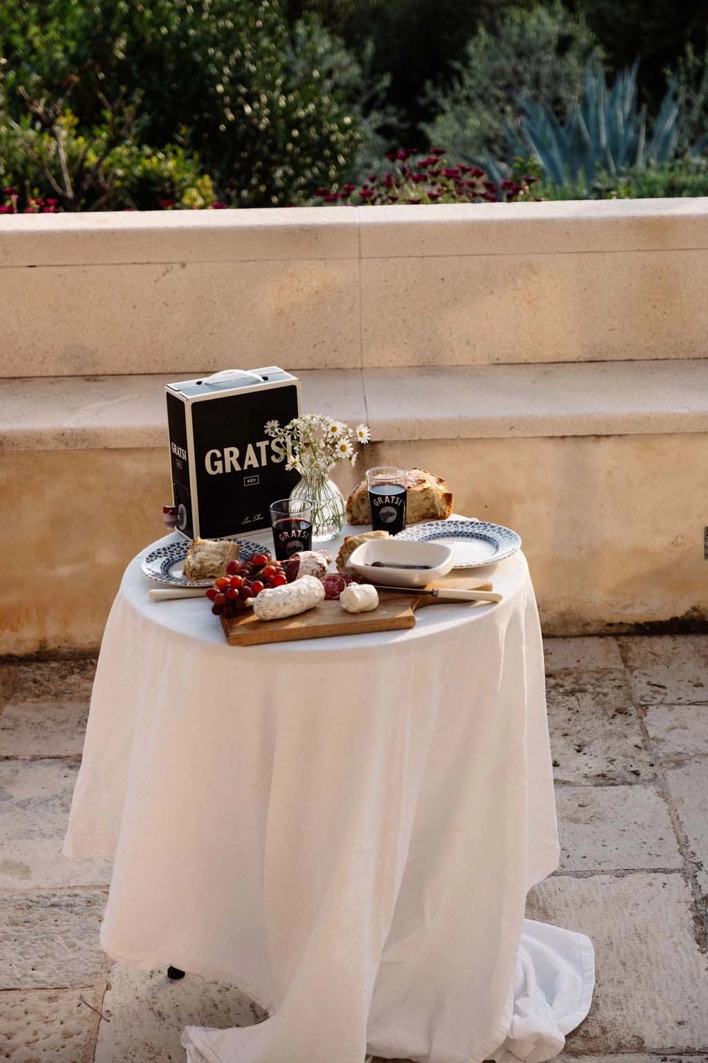 A round table covered with a white cloth displays cheese, bread, grapes, a box labeled Gratsi, and flowers in a vase, set outdoors on a stone patio with greenery in the background.
